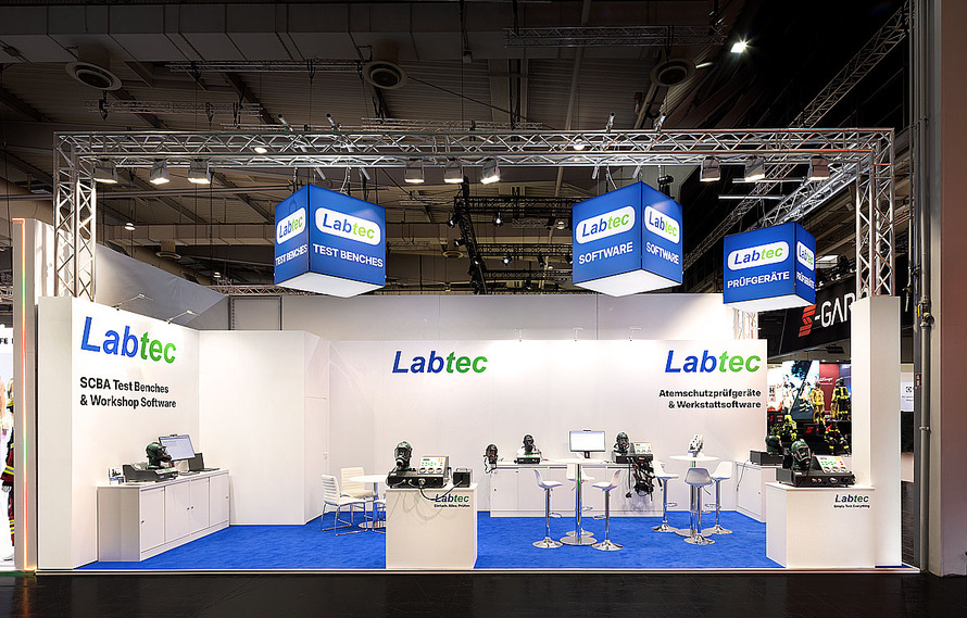 Messestand des Unternehmens Labtec in einer Messehalle. Der offen gestaltete Stand ist &uuml;berwiegend in Wei&szlig; und Blau gehalten und pr&auml;sentiert Pr&uuml;fger&auml;te sowie Softwarel&ouml;sungen f&uuml;r Atemschutztechnik. &Uuml;ber dem Stand h&auml;ngt eine silberne Traversenkonstruktion mit mehreren beleuchteten W&uuml;rfeln, auf denen die Begriffe &bdquo;Test Benches&ldquo;, &bdquo;Software&ldquo; und &bdquo;Pr&uuml;fger&auml;te&ldquo; zusammen mit dem Labtec-Logo zu sehen sind. Auf den wei&szlig;en R&uuml;ck- und Seitenw&auml;nden befindet sich mehrfach der Schriftzug &bdquo;Labtec&ldquo; sowie Hinweise auf Atemschutzpr&uuml;fger&auml;te, Werkstattsoftware und Pr&uuml;fst&auml;nde. Im Ausstellungsbereich stehen verschiedene Pr&uuml;fger&auml;te, Atemschutzmasken, Computerarbeitspl&auml;tze und Monitore auf wei&szlig;en Pr&auml;sentationstischen und Podesten. Mehrere Stehtische mit Barhockern und eine kleine Sitzgruppe dienen als Besprechungsbereiche. Der Boden des Messestandes ist mit einem auff&auml;lligen blauen Teppich ausgelegt.
