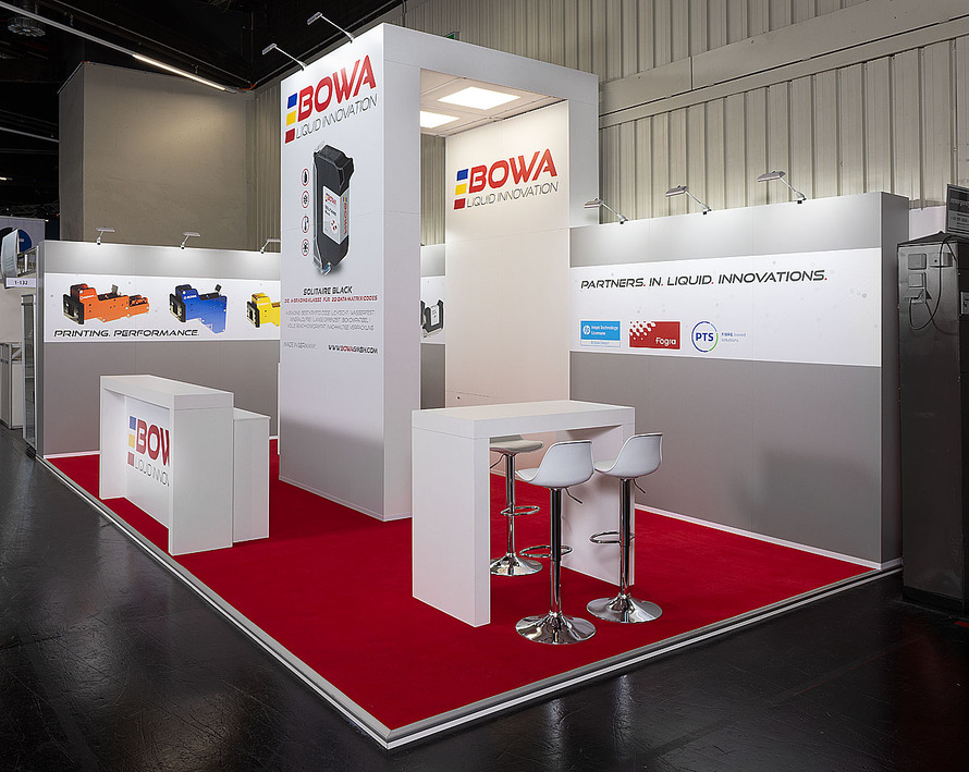 Messestand des Unternehmens BOWA auf einer Fachmesse. Der offen gestaltete Stand ist in Wei&szlig;, Grau und Rot gehalten. Ein roter Teppichboden bildet einen deutlichen Kontrast zu den hellen W&auml;nden und M&ouml;beln. Im Zentrum steht eine hohe, rechteckige S&auml;ule mit dem Firmenlogo &bdquo;BOWA Liquid Innovation&ldquo; sowie einer gro&szlig;formatigen Produktabbildung. Entlang der Standw&auml;nde werden verschiedene industrielle Kennzeichnungs- und Drucksysteme mit Produktgrafiken und technischen Informationen pr&auml;sentiert. Auf einer Wand ist der Slogan &bdquo;Partners in. Liquid. Innovations.&ldquo; zu lesen. Im vorderen Bereich befinden sich ein Empfangstresen sowie mehrere Besprechungstische mit wei&szlig;en Barhockern f&uuml;r Kundengespr&auml;che. Die klare, reduzierte Gestaltung legt den Fokus auf die pr&auml;sentierten Produkte und Informationen.