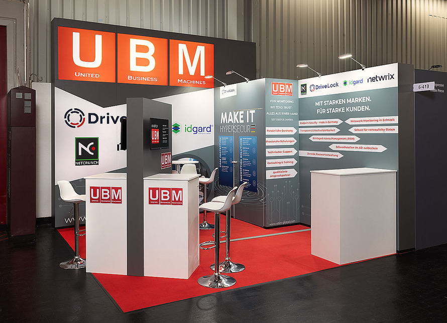 Messestand der UBM United Business Machines auf einer Fachmesse. Der kompakte Eckstand ist in den Farben Rot, Wei&szlig;, Grau und Schwarz gestaltet. Im oberen Bereich befindet sich eine gro&szlig;formatige Leuchtwerbung mit dem Logo &bdquo;UBM&ldquo; sowie dem ausgeschriebenen Unternehmensnamen &bdquo;United Business Machines&ldquo;. Die R&uuml;ck- und Seitenw&auml;nde pr&auml;sentieren verschiedene IT- und Cybersecurity-L&ouml;sungen sowie Partnerunternehmen. Sichtbar sind unter anderem Logos und Informationen zu den Bereichen Endpoint-Sicherheit, Netzwerk&uuml;berwachung, Privileged Access Management, Passwortverwaltung und Datenschutz. Im Zentrum des Standes steht eine hohe, freistehende Pr&auml;sentationss&auml;ule mit integriertem Bildschirm. Mehrere wei&szlig;e Theken und Stehtische mit Barhockern bieten Platz f&uuml;r Beratungsgespr&auml;che und Produktpr&auml;sentationen. Der rote Teppichboden hebt die Ausstellungsfl&auml;che deutlich vom dunklen Hallenboden ab. Die Gestaltung vermittelt einen klaren Fokus auf IT-Sicherheit, Monitoring und Infrastrukturmanagement.