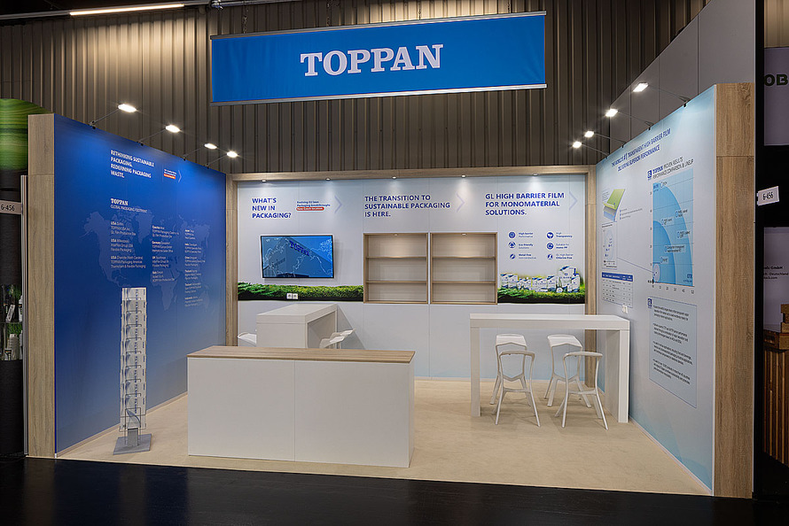 Messestand des Unternehmens TOPPAN auf einer Fachmesse. Der Stand ist offen gestaltet und kombiniert helle Holzoberfl&auml;chen mit wei&szlig;en M&ouml;beln sowie gro&szlig;fl&auml;chigen Informationsw&auml;nden in Blau und Wei&szlig;. &Uuml;ber dem Stand h&auml;ngt ein rechteckiges Banner mit dem Unternehmensnamen &bdquo;TOPPAN&ldquo;. Die R&uuml;ckwand pr&auml;sentiert Informationen zu nachhaltigen Verpackungsl&ouml;sungen, Monomaterial-Verpackungen und Barrierefolien. In der Mitte befindet sich ein Bildschirm zur Pr&auml;sentation digitaler Inhalte, daneben sind Produktabbildungen, Grafiken und technische Informationen zu sehen. Auf der linken Seite informiert eine gro&szlig;formatige Wand &uuml;ber internationale Standorte und Aktivit&auml;ten des Unternehmens. Im vorderen Bereich stehen ein Empfangstresen sowie ein Besprechungstisch mit mehreren St&uuml;hlen f&uuml;r Kundengespr&auml;che. Ein Prospektst&auml;nder erg&auml;nzt die Pr&auml;sentationsfl&auml;che. Die Gestaltung wirkt offen, &uuml;bersichtlich und auf die Vermittlung von Informationen zu Verpackungstechnologien und nachhaltigen L&ouml;sungen ausgerichtet.