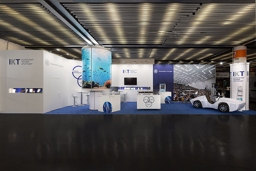 Gemeinschaftsstand des Instituts f&uuml;r Kunststofftechnik (IKT) der Universit&auml;t Stuttgart auf einer Fachmesse. Der weitl&auml;ufige Messestand ist &uuml;berwiegend in Wei&szlig; und Blau gestaltet und verf&uuml;gt &uuml;ber offene Pr&auml;sentations- und Gespr&auml;chsbereiche. Auf mehreren W&auml;nden sind die Logos des IKT sowie der Universit&auml;t Stuttgart angebracht. Im Zentrum des Standes befindet sich eine hohe, zylindrische S&auml;ule mit einem umlaufenden Motiv einer Unterwasserlandschaft mit Korallen und Fischen. Mehrere Empfangstheken und Informationsfl&auml;chen sind um diese S&auml;ule angeordnet. An den R&uuml;ckw&auml;nden werden Forschungsthemen, Projekte und Anwendungsbereiche der Kunststofftechnik pr&auml;sentiert. Ein Monitor dient zur Darstellung digitaler Inhalte. Auf der rechten Seite steht ein leichtes Forschungsfahrzeug mit offener Karosserie, das als Exponat ausgestellt wird. Blaue Teppichfl&auml;chen gliedern den Standbereich und schaffen einen Kontrast zum dunklen Hallenboden.