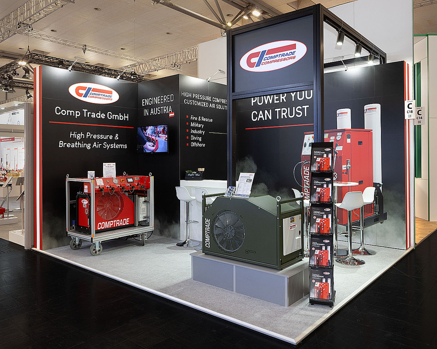 Messestand der Comp Trade GmbH auf einer Fachmesse. Der Stand ist in Schwarz, Wei&szlig; und Rot gestaltet und pr&auml;sentiert Hochdruck- und Atemluftsysteme. Gro&szlig;e schwarze Wandfl&auml;chen tragen das Firmenlogo sowie verschiedene Produkt- und Unternehmensinformationen. Zu lesen sind unter anderem Hinweise auf Hochdruckkompressoren und ma&szlig;geschneiderte Druckluftl&ouml;sungen f&uuml;r Feuerwehr, Rettungsdienste, Milit&auml;r, Industrie, Tauchsport und Offshore-Anwendungen. Im Ausstellungsbereich stehen mehrere technische Anlagen und Kompressorsysteme in Rot und Grau. Ein rotes mobiles Kompressorsystem befindet sich auf der linken Seite, w&auml;hrend im Vordergrund ein gr&ouml;&szlig;eres graues Aggregat auf einem Podest pr&auml;sentiert wird. Rechts sind weitere Kompressoranlagen sowie ein kleiner Besprechungsbereich mit wei&szlig;en Barhockern und einem Stehtisch zu sehen. Ein Prospektst&auml;nder mit Informationsmaterial steht an der Vorderkante des Messestandes.