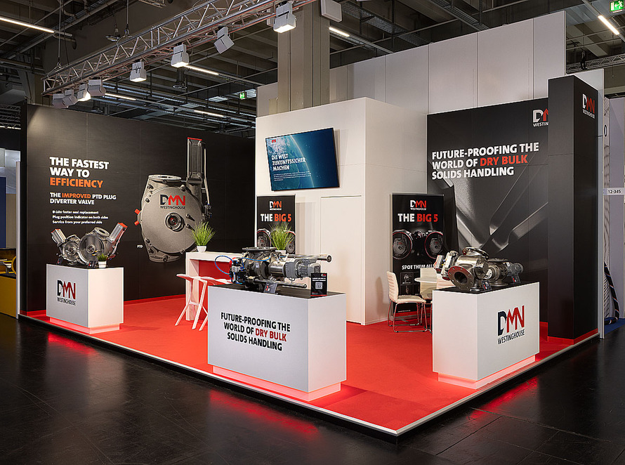 Messestand von DMN Westinghouse auf einer Fachmesse. Der offen gestaltete Eckstand ist in Schwarz, Wei&szlig; und Rot gehalten und pr&auml;sentiert technische Komponenten f&uuml;r die F&ouml;rderung und Verarbeitung von Sch&uuml;ttg&uuml;tern. Ein roter Teppichboden hebt die Ausstellungsfl&auml;che deutlich vom umliegenden Hallenboden ab. Auf mehreren wei&szlig;en Podesten sind gro&szlig;e industrielle Maschinen und Ventilsysteme aus Metall ausgestellt. Die schwarzen R&uuml;ckw&auml;nde zeigen gro&szlig;formatige Produktabbildungen sowie englischsprachige Informationen zu Effizienzsteigerung und L&ouml;sungen f&uuml;r die Sch&uuml;ttgutindustrie. Im Zentrum des Standes befindet sich ein Besprechungsbereich mit Tisch und Sitzgelegenheiten. Ein gro&szlig;er Bildschirm an einer wei&szlig;en Mittelwand zeigt digitale Inhalte des Unternehmens. &Uuml;ber dem Stand verl&auml;uft eine Traversenkonstruktion mit zahlreichen Scheinwerfern zur Ausleuchtung der Exponate.