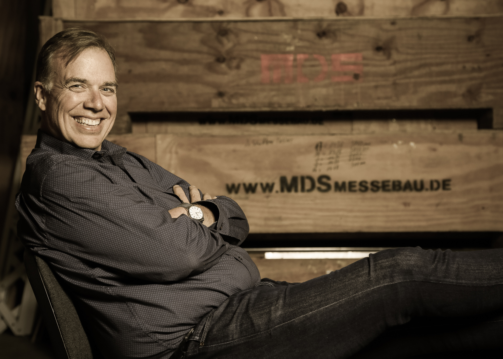 Foto des Geschäftsführers von MDS Messebau - Karsten Niemann Foto des Geschäftsführers von MDS Messebau - Karsten Niemann sitzend auf einer Kiste mit verschränkten Armen und einem sympathischen Lächeln