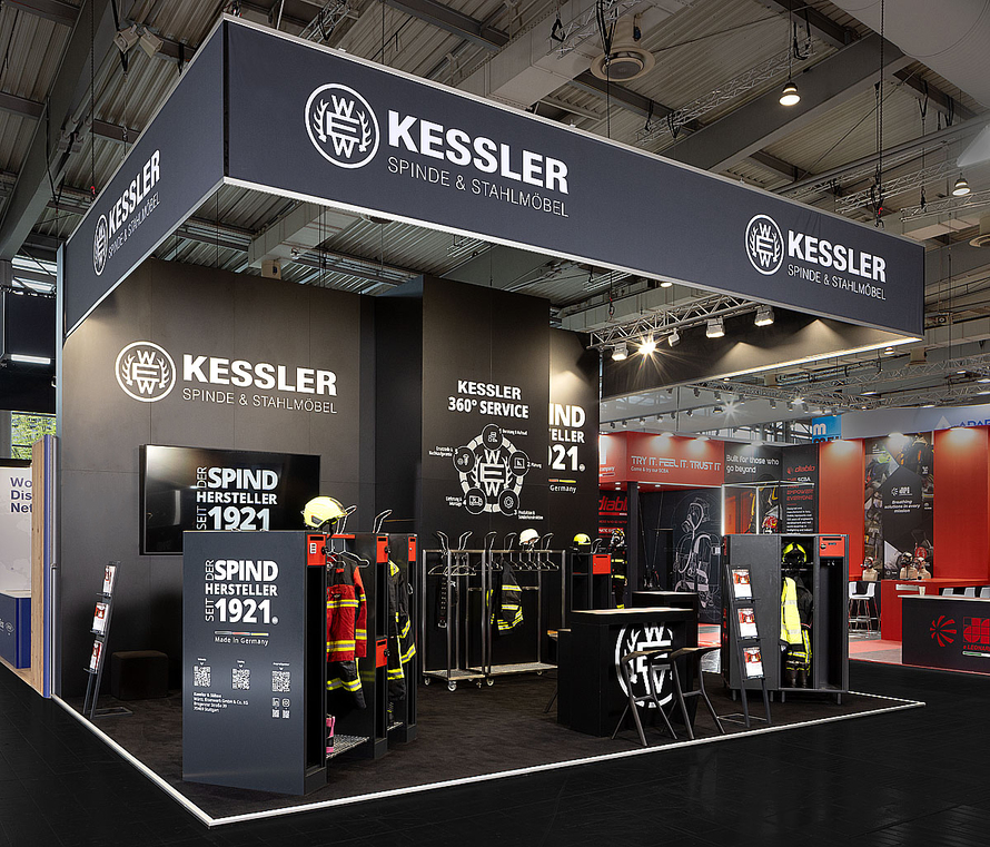 Messestand von Kessler Spinde & Stahlm&ouml;bel in einer Messehalle. Der offen gestaltete Stand ist in dunklen Grau- und Schwarzt&ouml;nen gehalten und wird von einer gro&szlig;en, abgeh&auml;ngten Beschilderung mit dem Firmenlogo sowie dem Schriftzug &bdquo;Kessler Spinde & Stahlm&ouml;bel&ldquo; umrahmt. Im Ausstellungsbereich sind verschiedene Stahlspinde und Garderobenschr&auml;nke pr&auml;sentiert. Einige Schr&auml;nke stehen ge&ouml;ffnet und zeigen Einsatzkleidung, Helme und Schutzausr&uuml;stung f&uuml;r Feuerwehr- und Rettungskr&auml;fte. Mehrere Kleiderst&auml;nder mit Einsatzjacken befinden sich im Zentrum des Standes. An den W&auml;nden sind gro&szlig;formatige Grafiken, Produktinformationen und Hinweise auf die Unternehmensgeschichte seit 1921 zu sehen. Im Vordergrund befinden sich ein Beratungstresen, Sitzgelegenheiten und Informationsst&auml;nder.