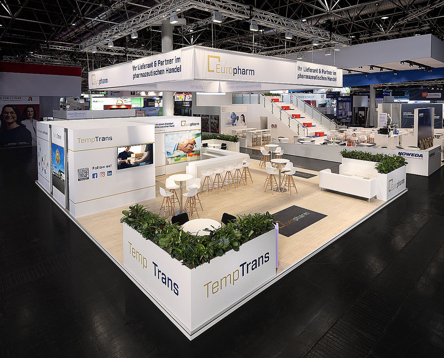 Gro&szlig;z&uuml;giger Messestand von Europharm auf einer Fachmesse. Der offen gestaltete Stand ist &uuml;berwiegend in Wei&szlig; gehalten und verf&uuml;gt &uuml;ber helle Bodenfl&auml;chen, moderne Sitzgelegenheiten sowie mehrere Besprechungs- und Aufenthaltsbereiche. &Uuml;ber dem Stand h&auml;ngt eine gro&szlig;fl&auml;chige, beleuchtete Deckenkonstruktion mit dem Europharm-Logo und dem Schriftzug &bdquo;Ihr Lieferant & Partner im pharmazeutischen Handel&ldquo;. Im Zentrum des Standes befinden sich lange Theken, Stehtische und zahlreiche Sitzpl&auml;tze f&uuml;r Gespr&auml;che mit Besucherinnen und Besuchern. Eine breite Treppe mit roten Stufen f&uuml;hrt zu einer erh&ouml;hten Ebene im hinteren Bereich des Messestandes. Mehrere Informationsfl&auml;chen, Bildschirme und Unternehmensdarstellungen pr&auml;sentieren Leistungen und Angebote aus dem pharmazeutischen Handel. Begr&uuml;nte Pflanzenelemente und Lounge-M&ouml;bel schaffen eine offene und einladende Atmosph&auml;re.